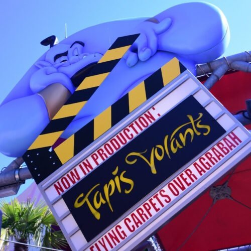 Geest Genie Tapis Volants Blauwe geest Genie met een filmklapper met daarop in gele letters Tapis Volants bij de entree van de attractie